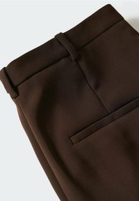 Pantalon marron foncé sur mesure avec passants pour ceinture et une seule poche arrière horizontale visible sur un fond clair.