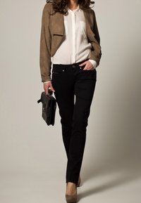 Ruskea mokkanahkainen lyhyt takki, valkoinen napitettava paita, mustat kapeat housut, beige korkokengät, musta nahkalaukku käsissä, neutraali tausta.