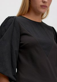 Vrouw in een zwart jurkje met gestructureerde pofmouwen en een driehoekig paneel op de borst, afgebeeld van nek tot halverwege de torso tegen een effen achtergrond.