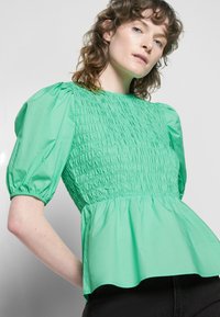 Top verde de mangas bufantes feito de tecido franzido, com corpete pregueado e bainha rodada, tendo uma textura suave e decote redondo.