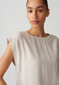 Helles Beige, ärmelloses Oberteil aus weichem Stoff mit Bootsausschnitt und subtiler Textur. Verfügt über breite Armausschnitte und eine lockere Passform.