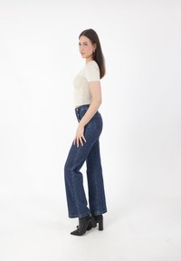 Kobieta z ciemnymi włosami stoi z boku, ubrana w beżową bluzkę z krótkim rękawem, niebieskie dżinsy z wysokim stanem oraz czarne botki na obcasie.