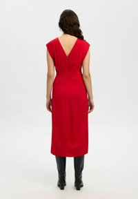 Frau mit langen dunklen Haaren, die ein ärmelloses rotes Midi-Kleid mit V-ausschnitt und schwarze knielange Stiefel trägt, steht vor einem schlichten Hintergrund.