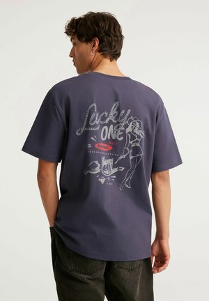 Mężczyzna z krótkimi brązowymi włosami i kolczykiem nosi ciemnofioletową koszulkę z napisem "Lucky One" i grafiką kobiety w stylu pin-up na plecach.