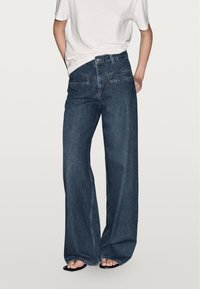 Breda ben i mörk denimjeans med framfickor, hög midja och synlig sömnad. Kombinerat med en enkel vit kortärmad skjorta.