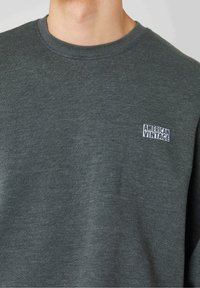 Sweat-shirt à col rond gris foncé en tissu doux, avec un petit logo blanc "American Vintage" sur la poitrine.