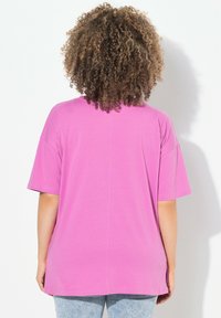 Camiseta rosa de gran tamaño con mangas cortas, fabricada en un material transirable y con un dobladillo redondeado. Presenta una costura central como detalle de diseño adicional.