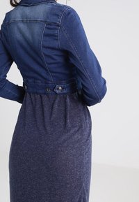 Jeansjacka med mörkblå tvätt, figurnära kropp och knappdetaljer; bärs över en mjuk, marinblå klänning med gathered midja.