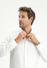 Witte overhemdd met een kraag, lange mouwen en knoopdetails, wordt vastgemaakt getoond. Gladde stof, getailleerde pasvorm, minimalistisch ontwerp.