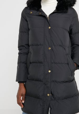 Long manteau d'hiver matelassé noir avec des boutons dorés, un col en fourrure noire et des poches latérales, porté sur un pull à col roulé blanc.