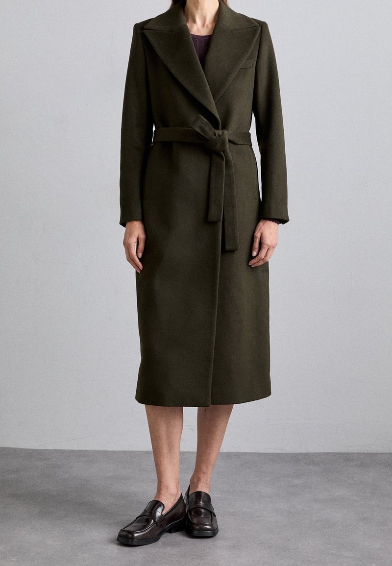 Cappotto di lana verde oliva con vita rifinita da cintura, maniche lunghe e colletto a rever. Tessuto liscio, lunghezza al ginocchio e dettagli minimi.