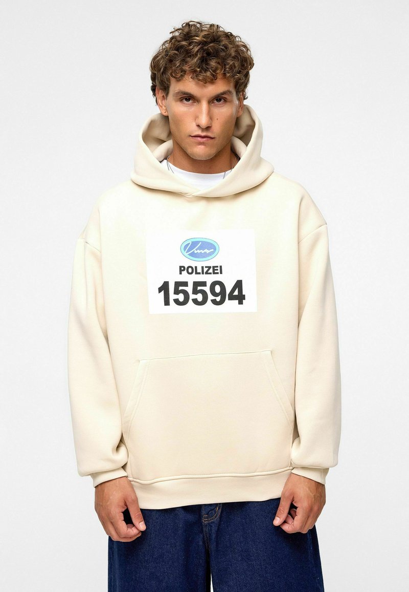 Crème hoodie met een voorkantoor, met een grote afdruk van "POLIZEI 15594" in zwart en blauw. De stof lijkt zacht en gestructureerd.