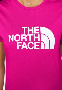 Camiseta de algodón rosa con una gran impresión blanca de "THE NORTH FACE" y un logo curvado en forma de media luna. Diseño clásico de cuello redondo.