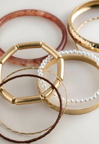 Five bangles in varying materials: two brown-toned, one golden with a textured finish, a pearl-accented design, and a slender gold wire.