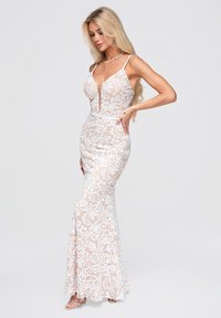 Femme aux longs cheveux blonds portant une robe beige longue jusqu'au sol avec de la dentelle florale blanche, fines bretelles et un profond décolleté en V, posant avec une main sur la hanche.