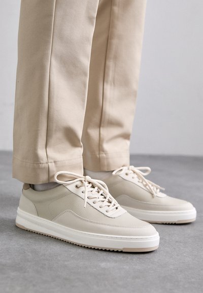 Beige sneakers met een textuur bovenkant, witte zolen en gestikte accenten, gecombineerd met beige broeken, tentoongesteld op een grijze ondergrond.