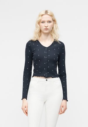 Ξανθιά γυναίκα που φορά μπλε σκούρο crop top με μακριά μανίκια, κουμπιά και λευκά πουά, καθώς και λευκό παντελόνι με ψηλή μέση, στέκεται μπροστά σε λευκό φόντο.