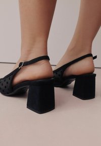 Mustat slingback-kengät, joissa on pilkullinen kuvio, neliönmuotoiset korot ja teksturoitu kangas. Varustettu säädettävällä remmillä ja sileällä sisävuorella.