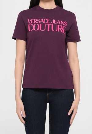 Persona che indossa una t-shirt viola a maniche corte con testo rosa "VERSACE JEANS COUTURE" e jeans blu scuro su uno sfondo semplice.