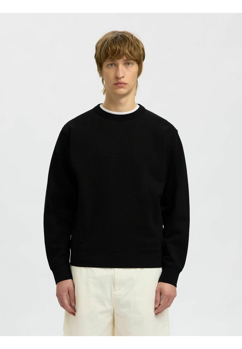 Sudadera negra de manga larga y cuello redondo. Textura suave, corte holgado y puños acanalados. Llevada con pantalones de color crema.