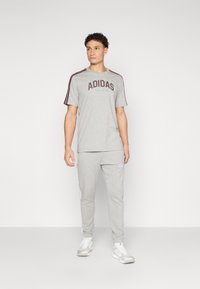 Pelēks Adidas T-krekls ar logotipu un ušakānu akcentiem, apvienots ar atbilstošām sporta biksēm ar baltām sānu svītrām. Apavi ir balti.