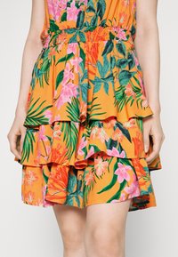 Femme portant une jupe orange à étages avec des feuilles vertes et des motifs floraux roses, mains détendues le long du corps.