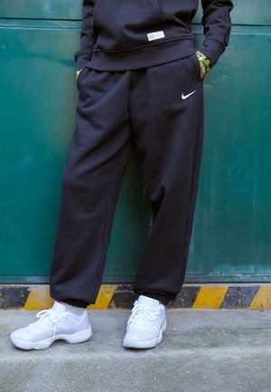Personne portant un pantalon de survêtement Nike noir et des baskets blanches, debout contre un mur métallique vert, les mains dans les poches.