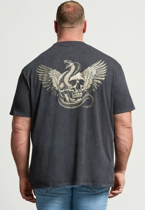 Mand iført mørkegrå T-shirt med stort vinget kranie og snoet slange grafik på ryggen, kombineret med lyseblå jeans.