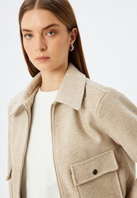 Giacca beige con zip realizzata in tessuto testurizzato, con colletto a punta e una tasca laterale con dettaglio a bottone a pressione. Camicia bianca sotto.