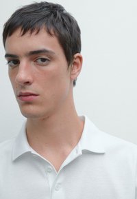Bílá polo tričko s měkkou texturou, s klasickým límečkem a dvěma knoflíky u krku, nošeno osobou, která se dívá přímo do kamery.