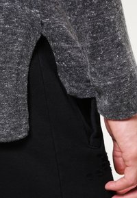 Grijze gebreide trui met een textuurafwerking en een licht gebogen zoom, gecombineerd met zwarte broek met een zijzakdetail.