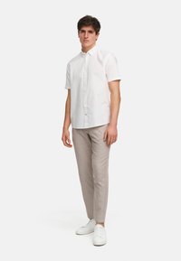 Camisa blanca de manga corta con botones, combinada con pantalones ajustados beige claros y zapatillas blancas, de pie contra un fondo liso.