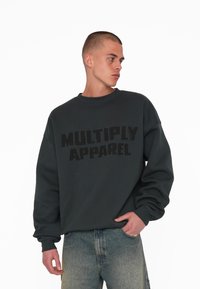 Sweatshirt gris foncé avec le texte noir en gras "MULTIPLY APPAREL". Il présente un col rond et une coupe décontractée avec des poignets aux manches.