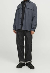 Mörkgrå knapp-up skjorta med två bröstfickor, bärs över en svart skjorta med hög krage, i kombination med mörka denimbyxor och svarta skor.