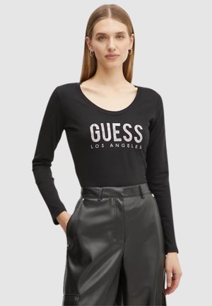Maglietta nera a maniche lunghe con scollatura rotonda e logo argento "GUESS LOS ANGELES" sulla parte anteriore. Indossata con pantaloni neri a vita alta.