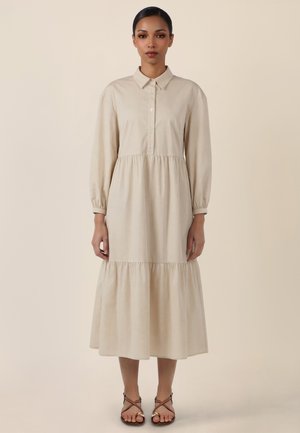 Woman standing wearing a long-sleeve beige midi dress with a collar, buttoned front, gathered tiers, and black strappy flat sandals.