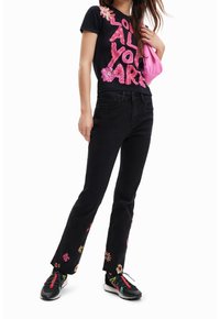 Černé tričko s růžovým květinovým potiskem a textem, doplněné černými vyšívanými džínami s květinovými vzory a barevnými sportovními botami.