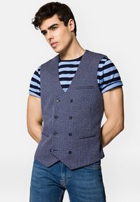 Kamizelka w deseń houndstooth w kolorze granatowym i białym, z dekoltem w serek, podwójnymi guzikami i kieszenią z przodu, nałożona na niebieską koszulkę w paski granatowe.