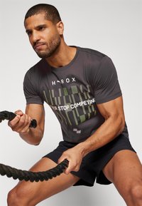 Camiseta atlética de color gris oscuro con texto gráfico verde, combinada con pantalones cortos negros. La persona está haciendo ejercicio con una cuerda gruesa y negra.