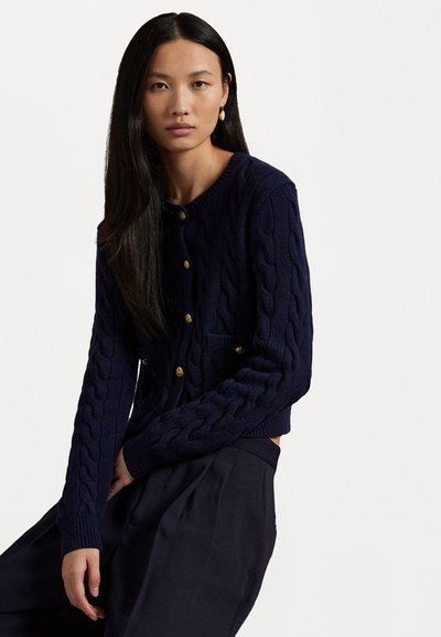 Laivaston sininen palmikkoneuleinen kardigan, jossa on pyöreä pääntie, kultaiset napit ja kaksi etutaskua; valmistettu teksturoidusta kankaasta lämmön takaamiseksi.