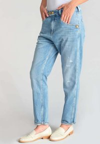 Jean en tissu denim bleu clair avec une coupe décontractée, présentant deux poches avant, des boutons en laiton et des détails usés. Associé à des mocassins blancs cassés.