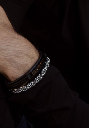 Black leather bracelet with silver chain detail, featuring a smooth texture and a rectangular clasp, worn on a wrist with light hair.