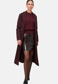 Cappotto lungo bordeaux sopra un maglione bordeaux a trama e una mini gonna in faux pelle lucida marrone, abbinato a mocassini neri e collant trasparenti.