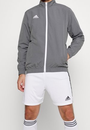 Mann trägt eine graue Adidas Zip-Up-Jacke und weiße Adidas-Shorts mit gestreiften Socken, steht mit einer Hand in der Tasche.
