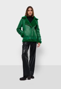 Zelená kožuchová kožená bunda s klopeným golierom, opaskom v páse a zipsami. Nosená s čiernymi koženými nohavicami a širokými podpätkami.