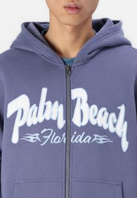 Lilla zip-up hoodie med "Palm Beach Florida" i store hvide bogstaver. Har en hætte med snor og kængurulomme foran.