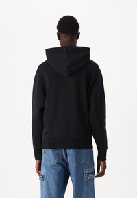 Un sweat à capuche noir avec une grande capuche, des poignets et un ourlet côtelés, porté sur un jean bleu clair retroussé. Design ajusté, texture lisse et détails minimalistes.