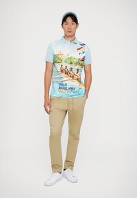 Πολάκι με μπλε γραδίαντ και σκηνή κωπηλασίας, κοντά μανίκια και γιακά; συνδυασμένο με ανοιχτό καφέ παντελόνι και λευκά sneakers.