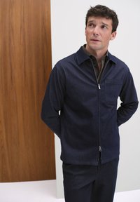 Marinblå denimjacka med dragkedja fram och krage, med en bröstficka och långa ärmar, ihop med matchande mörka byxor.