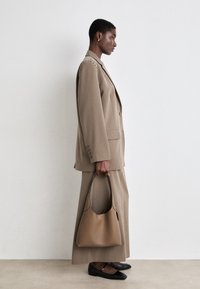 Femme en tailleur beige sur mesure tenant un sac à main en cuir marron clair, portant des ballerines noires, debout de profil contre un mur blanc uni.